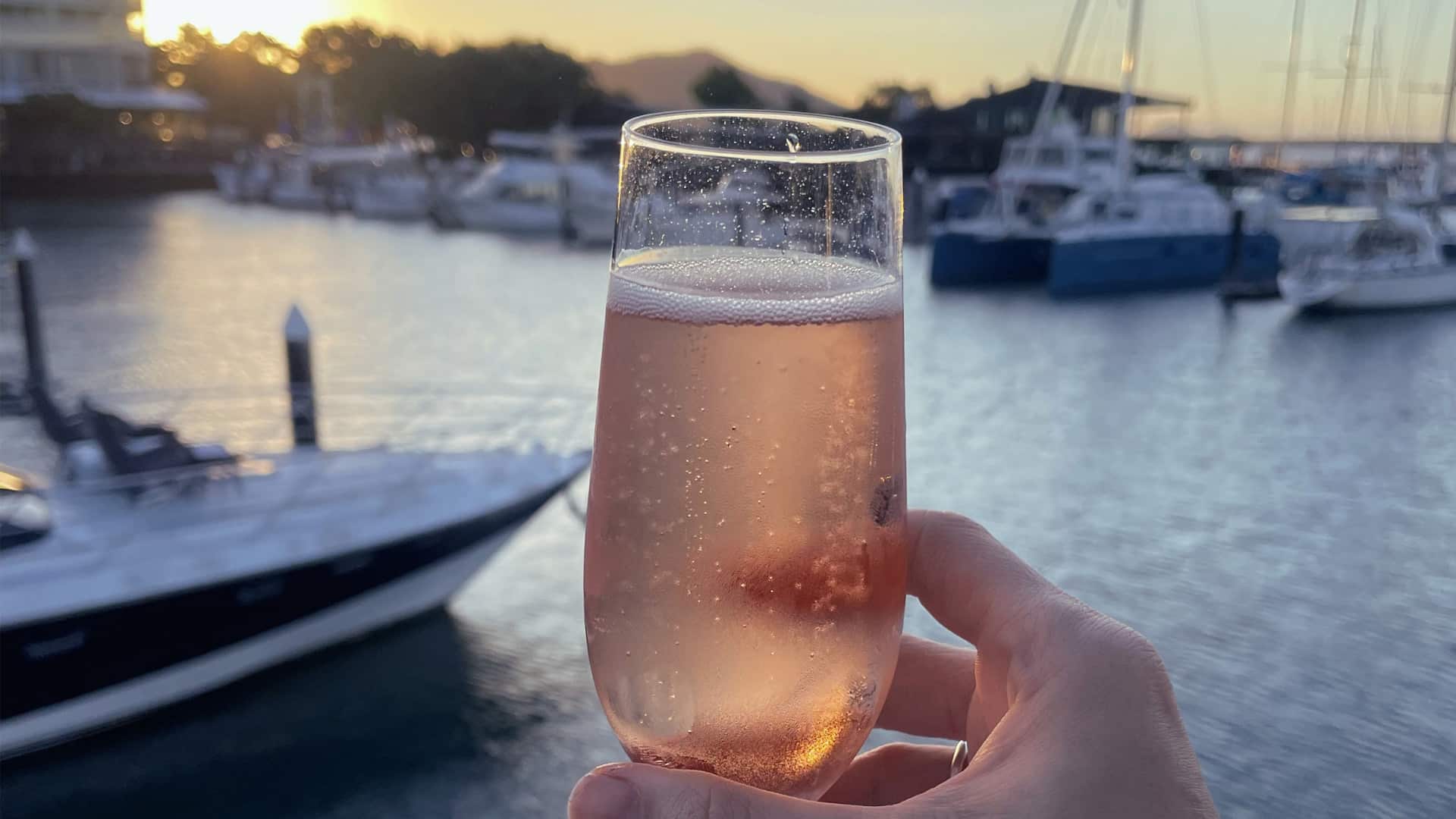 Pink sparkling rosé in a flute held up at a marina sunset with boats in the background warm glow on the water