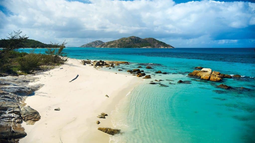 White-sand tropical beach with clear turquoise water, rocks along the shore, and a distant hilly island under a partly cloudy sky.