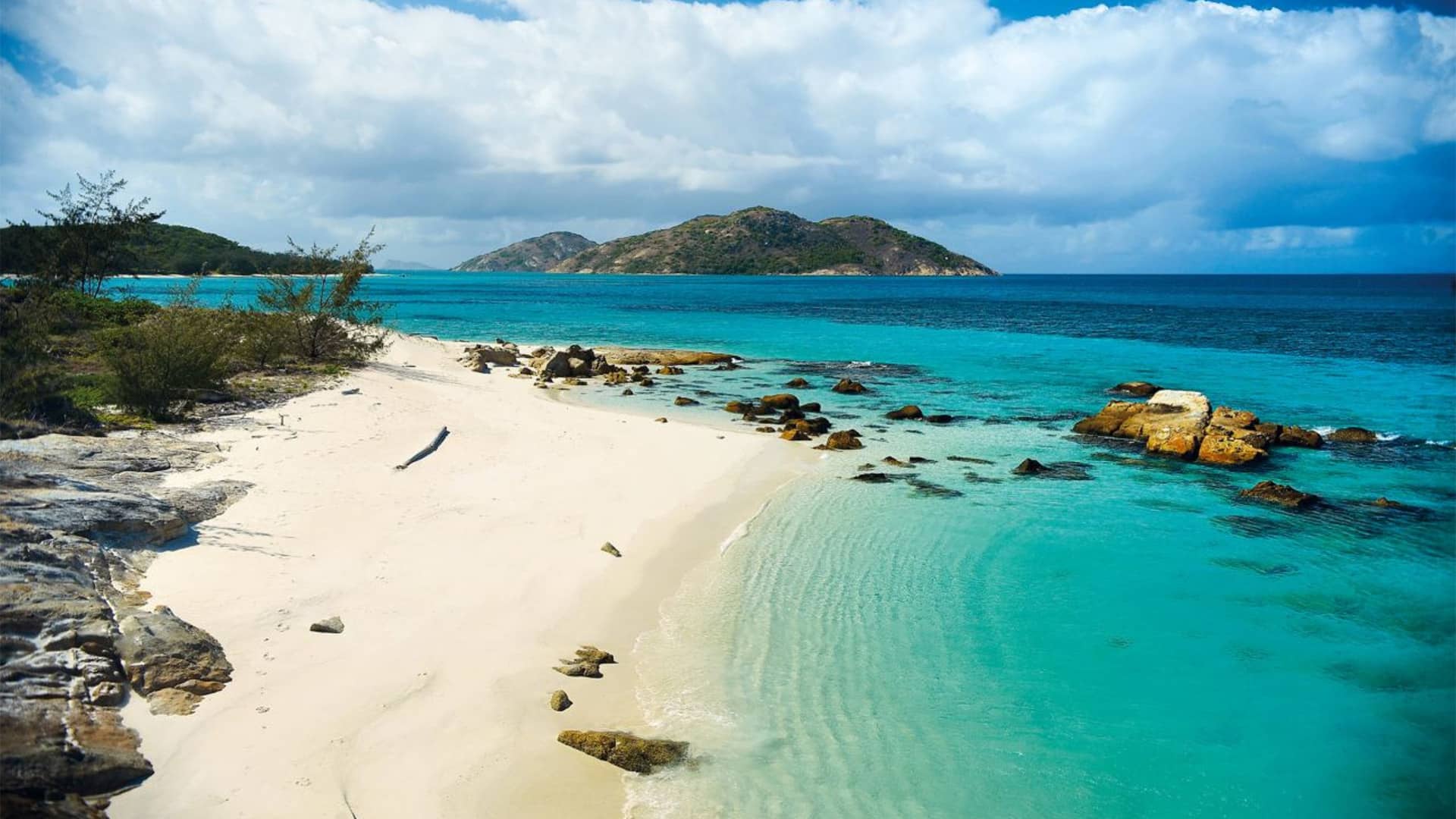 White sand tropical beach with clear turquoise water rocks along the shore and a distant hilly island under a partly cloudy sky