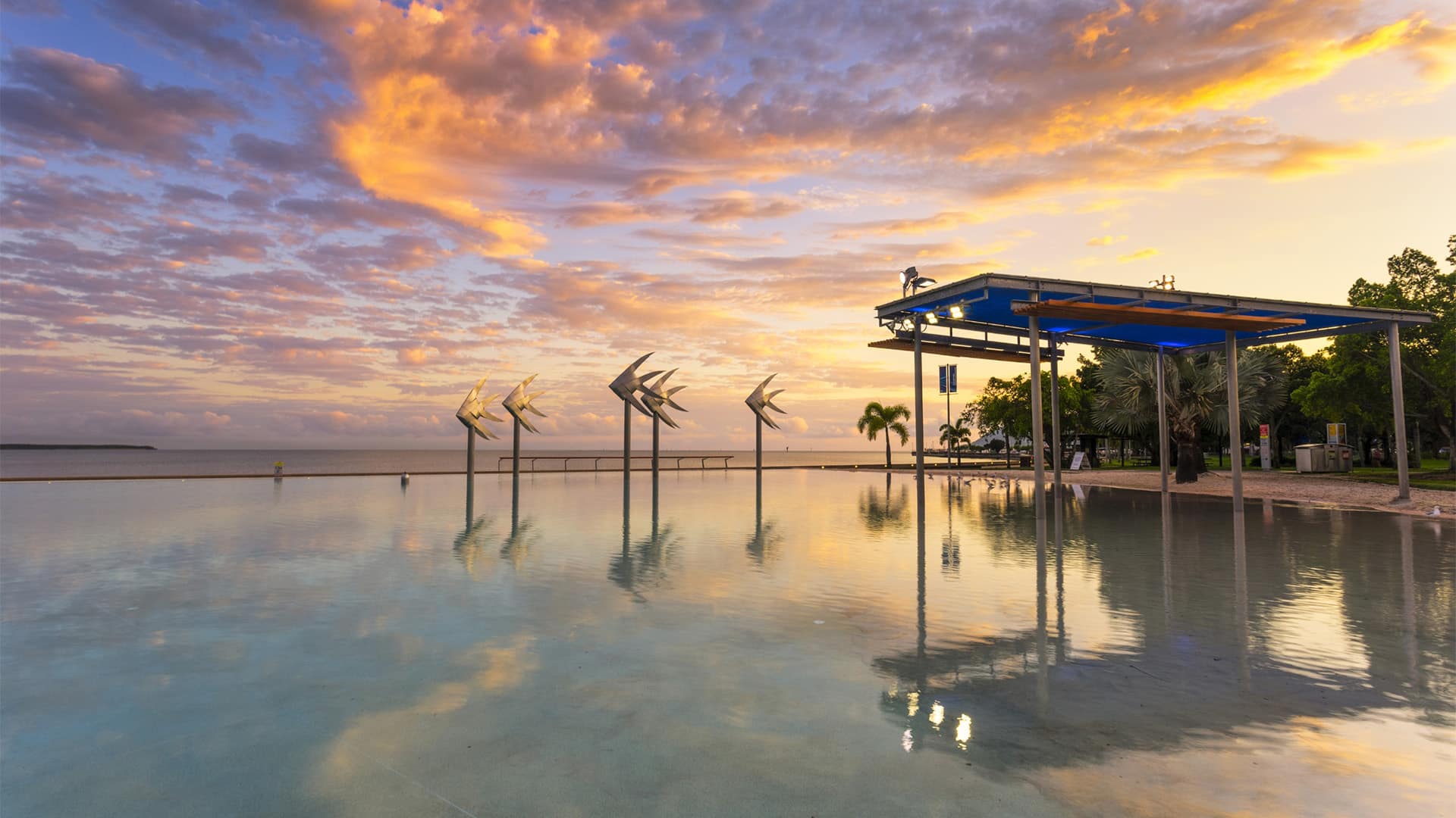 Sunset over a calm pool with angled metal wind sculptures and a blue shaded pavilion by the shore